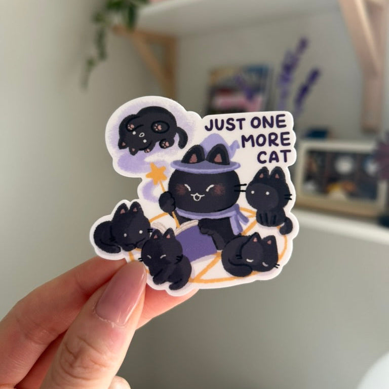 Sticker with cartoon cats and text 'Just one more cat' held by a hand, with a blurred indoor background.